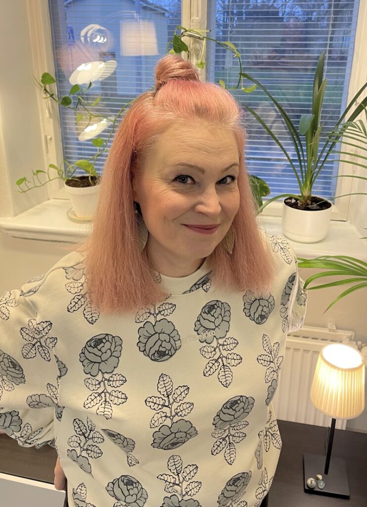 Psykoterapeutti Heli Tyrkko katsoo hymyillen kameraan vastaanottohuoneessaan. Psykoterapia Juurille Oy, Hämeenlinna.